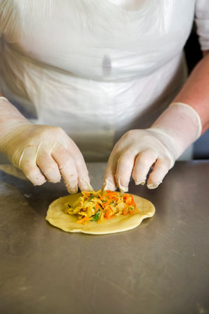 A chef is working in a kitchen. Wearing gloves, the chef adds a filling made of vegetables to a...