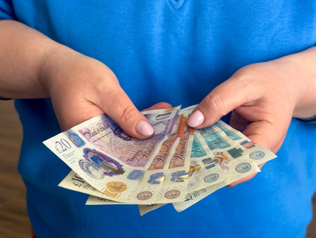 A person holds several British pound sterling notes in both hands. The notes show various colors and...