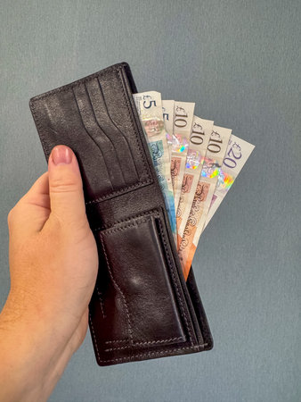 A person holds a black wallet open, displaying British pound notes in different amounts. The notes...