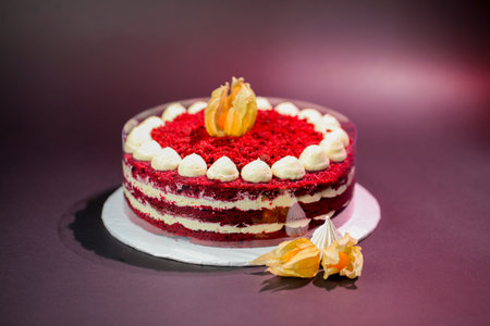 A red velvet cake with layers of cream stands on a white base. Decorative fruit is placed around the...