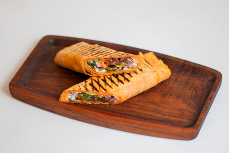 A wrap filled with meat and vegetables sits on a wooden plate. It is cut in half to show the...