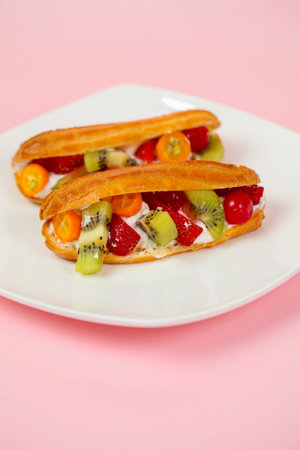 Two eclairs filled with fresh fruits sit on a white plate. The background is pink. The fruits...