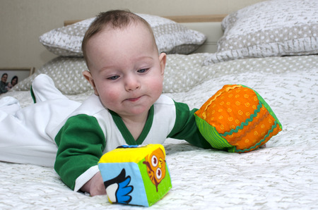 Little boy lying on his stomach on a white bed with toys in the hands.の写真素材