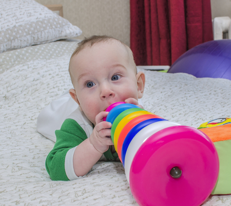 Little boy in a green suit lying on a white bed and bites color toys.の写真素材