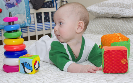 Boy toddler is lying on a white bed with toys of different colors looking to the left.の写真素材