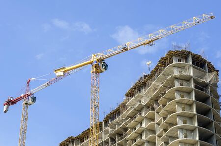 Yellow and red construction cranes against a blue sky and a gray house under construction.の写真素材