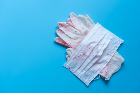 Close up surgical mask and latex gloves with blood stain on blue background. Used surgical masks for...