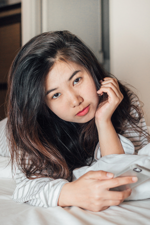 Pretty young asia woman holding cell phone and looking at camera on the bedの写真素材