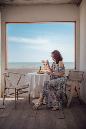 A woman traveler sits by the window relaxing and enjoying her vacation at the sea cafe.の写真素材