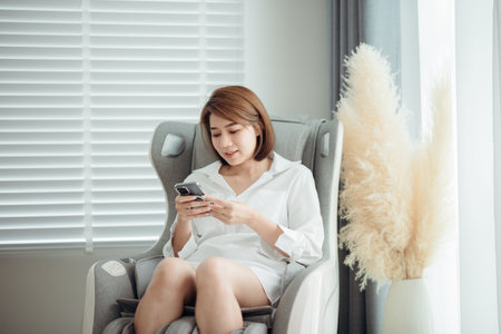 A woman relaxing and using her mobile phone and sitting on the massage chair in the living room.の写真素材
