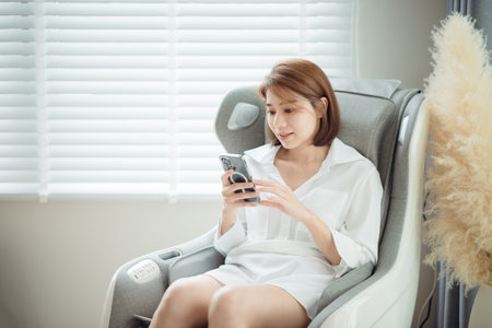 A woman relaxing and using her mobile phone and sitting on the massage chair in the living room.の写真素材