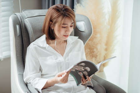 A woman reading a book and listening to music while sitting on the massage chair in the living room.の写真素材