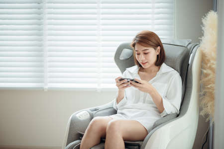 A woman relaxing and playing games on her mobile phone and sitting on the massage chair in the living room.の写真素材