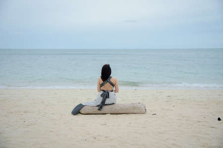 Rearview of a lonely woman sitting on the log and looking to the sea.の写真素材