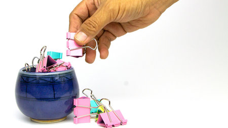 Close up shot of hand picking up colorful paperclips from blue ceramic container on white background with copy space.の写真素材