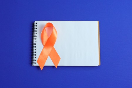Orange ribbon and open notebook on blue background, healthcare and medicine concept. Multiple Sclerosis awareness. Leukemia awareness. Kidney cancer awareness.の写真素材