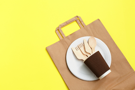 Eco-friendly disposable utensils made of bamboo wood on paper bag on yellow background. Draped spoons, fork, knives, bamboo bowls with paper cups and packet. Concept of disposable tableware.の写真素材