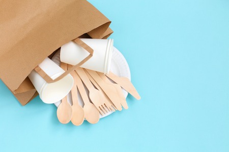 Eco-friendly disposable utensils made of bamboo wood in paper bag on blue background. Draped spoons, fork, knives, bamboo bowls with paper cups and packet. Concept of disposable tablewareの写真素材