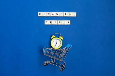 Phrase Financial Crisis and shopping trolley with alarm clock on blue background. Financial crisis...