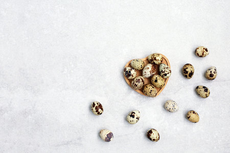 Small quail eggs in wooden bowl in the shape of a heart on light stone background with. Eco...