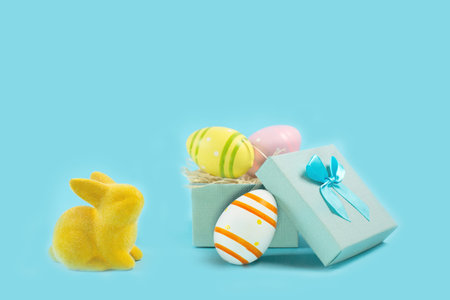 Yellow Easter bunny rabbit with painted egg in gift box on blue background.