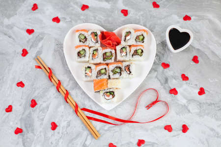 Sushi rolls with salmon on white heart-shaped plate and wooden sushi sticks among red confetti heartsの写真素材