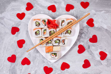 Sushi rolls with salmon on white heart-shaped plate and wooden sushi sticks among red confetti heartsの写真素材