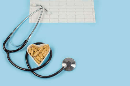 Herbal pills in heart shaped wooden plate, stethoscope and cardiogram on blue background.の写真素材