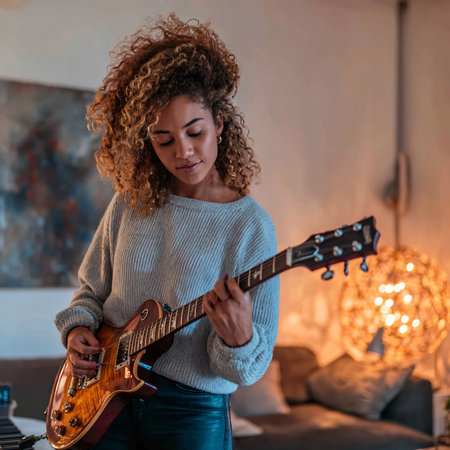 Young mixed-race woman with curly hair playing electric guitar in a cozy living room.の素材