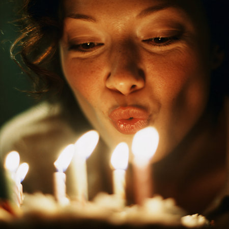 A young woman with curly brown hair blows out candles on a birthday cake.の素材
