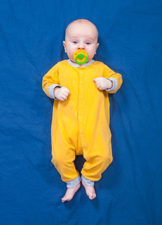 Three month baby boy in a yellow slip with pacifier lies on a blue cloth background
