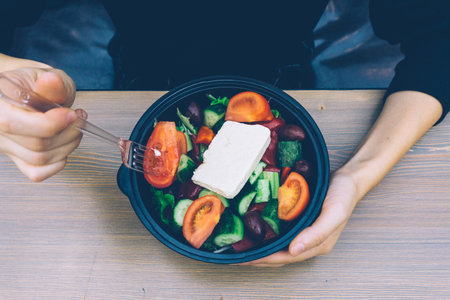Fast food Greek salad with fresh vegetables, feta cheese and olives on wooden table with female hands.の写真素材