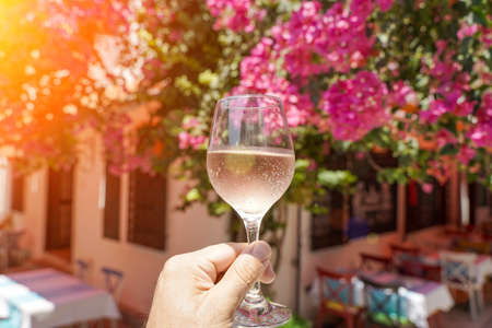 Glass of wine in hand. A glass of young fresh rose wine against the backdrop of a summer cafe in a Mediterranean seaside tourist town in the summer under sunlight. Summer, travel, lifestyle, relaxation, and enjoyment concept. high quality photoの写真素材