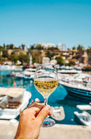 Glass of wine in hand. A glass of white wine against the backdrop of the Mediterranean sea and the port with yachts in a tourist town in the summer under sunlight. Summer, travel, lifestyle, relaxation, and enjoyment concept. high quality photoの写真素材