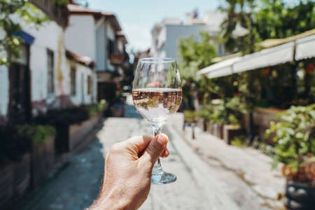 Glass of wine in hand. A glass of young fresh rose wine against the backdrop of a summer cafe in a Mediterranean seaside tourist town in the summer under sunlight. Summer, travel, lifestyle, relaxation, and enjoyment concept. high quality photoの写真素材
