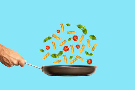 Hand with a frying pan and italian pasta with tomatoes and basil on a colored blue background. Food collage, minimal art creative concept.の写真素材