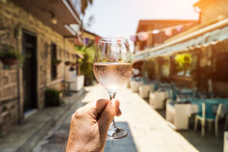 Glass of wine in hand. A glass of young fresh rose wine against the backdrop of a summer cafe in a Mediterranean seaside tourist town in the summer under sunlight. Summer, travel, lifestyle, relaxation, and enjoyment concept. high quality photoの写真素材