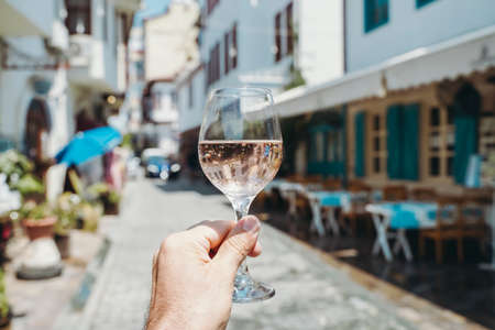 Glass of wine in hand. A glass of young fresh rose wine against the backdrop of a summer cafe in a Mediterranean seaside tourist town in the summer under sunlight. Summer, travel, lifestyle, relaxation, and enjoyment concept. High quality photoの写真素材