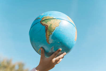 Hand with a globe on a clear sky background. Childrens hand with earth planet globe in sunny summer weather. Environmental protection, ecology, nature concept.の写真素材