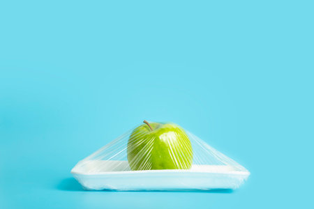 Fruits in plastic packaging from the supermarket are minimal. Apple in cellophane and non-degradable plastic on a blue background. Biodegradable product packaging, environmental protection, nature pollution. High quality photoの写真素材