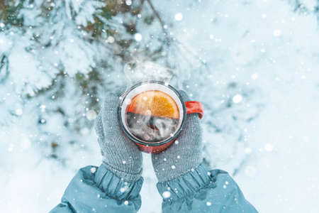Mulled wine in the hands of a girl during a snowfall in the forest. Winter hot drinks with aromatic spices of cinnamon, cardamom and orange. Warmth, comfort and atmosphere of December nature and Christmas. High quality photoの写真素材