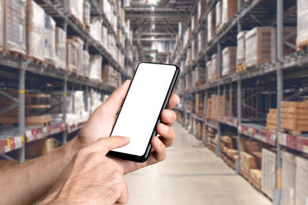 Warehouse with a hand holding an empty smartphone. Blurred industrial warehouse with goods and products. Logistics, business, delivery, services conceptの写真素材