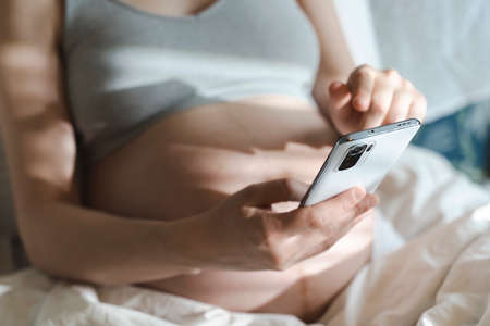 A pregnant woman uses a smartphone while relaxing in her home bed. Internet, shopping, communication during pregnancy and before the birth of a child. Womens and newborn healthの写真素材