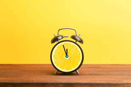Alarm clock with citrus fruit concept. Lemon or orange and clock on empty color background. Vitamins, proper nutrition, health, detox, diet concept.の写真素材