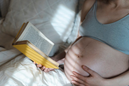 A pregnant woman reads a book and strokes her belly with a baby before childbirth while relaxing in a sunny bright bedroom lying on a bed. Selected focus. Women's health, pregnancy, conception, childbirth concept. High quality photoの写真素材