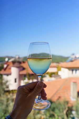 Wine drink in a glass in hand. White fresh wine in a transparent glass against the backdrop of the nature of the village in the summer on a sunny day. Drink, good mood, alcohol concept.の写真素材