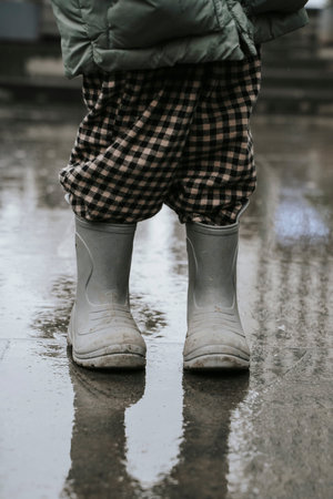 Toddler in Grey Rubber Boots and Plaid Pants on Wet Rainy Streetの写真素材