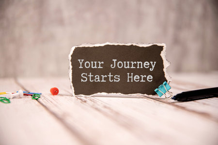 Closeup businesswoman hands holding white card sign with Your Journey Starts Here text message isolated on gray wall office background. Retro instagram style imageの写真素材