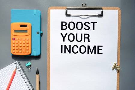 A hand holding a pencil over the text Boost your income on yellow sticky paper on a black background.の写真素材