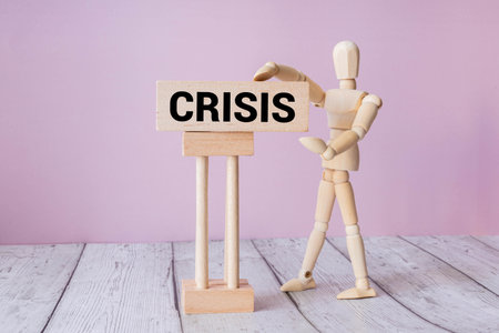 A wooden mannequin stands next to a wooden sign displaying the word Crisis against a pink background.の写真素材